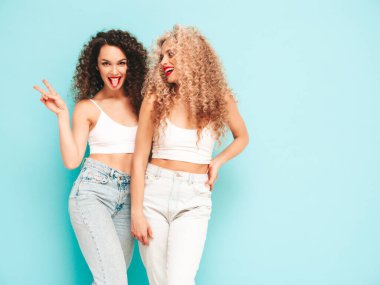 Yaz kıyafetleri içinde gülümseyen iki güzel hippi kadın. Saç stili kıvırcık saçlı moda modeller stüdyoda eğleniyor. Mavide izole edilmiş seksi bir kadın. Mutlu ve neşeli. Güneş gözlükleriyle