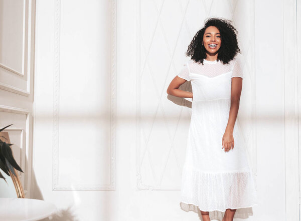 Portrait of beautiful black woman with afro curls hairstyle. Smiling model dressed in white summer dress. Sexy carefree female posing near wall in studio. Tanned and cheerful. At sunny day