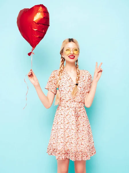 年轻美丽的微笑女性穿着时髦的红色衣服 一个性感无忧无虑的金发女人在工作室的蓝色墙壁附近摆姿势 有乐趣的正面模型 在太阳镜里快乐快乐 拿着心形气球 — 图库照片
