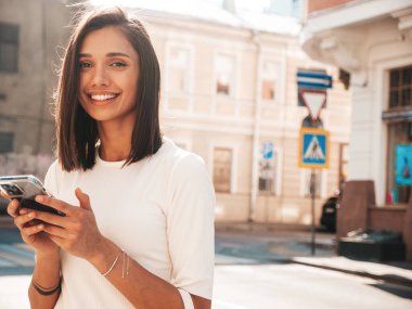 Modayı takip eden yaz elbisesi giymiş güzel, gülümseyen hippi kadın. Günbatımında sokakta poz veren kaygısız seksi kadın. Pozitif model. Akıllı telefon uygulamaları kullanarak. Cep telefonu ekranına bakıyorum.
