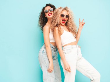 Yaz kıyafetleri içinde gülümseyen iki güzel hippi kadın. Saç stili kıvırcık saçlı moda modeller stüdyoda eğleniyor. Mavide izole edilmiş seksi bir kadın. Mutlu ve neşeli. Güneş gözlükleriyle