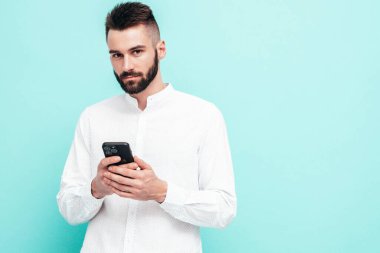 Yakışıklı, kendine güvenen, seksi, kotlu ve gömlekli bir adam. Moda hippi erkek stüdyoda mavi duvarın yanında poz veriyor. Akıllı telefonu tutuyorsun. Cep telefonu ekranına bakıyorum. Uygulamalar kullanılıyor