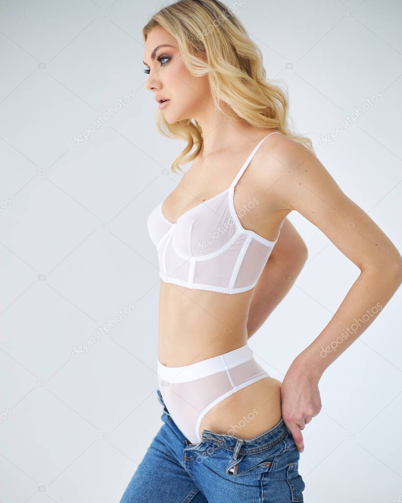 Beautiful girl is putting in jeans. Photo of a perfect body in  underwear on a white background. Diet, body care, fitness, skin and beauty concept. Pretty female model with athletic body, curly hair wearing white lace lingerie enjoying sunny morning.