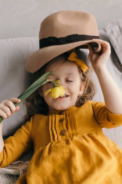 Sarı elbiseli küçük kız gözleri kapalı gülümsüyor ve narsist çiçekleri kokluyor.