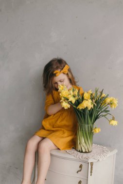 Sarı elbiseli, güzel bir fiyonklu küçük kız elinde çiçekle barok bir komodinin üzerinde oturuyor.