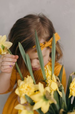 Sarı elbiseli, güzel fiyonklu küçük kız elinde narsist bir çiçek tutuyor.