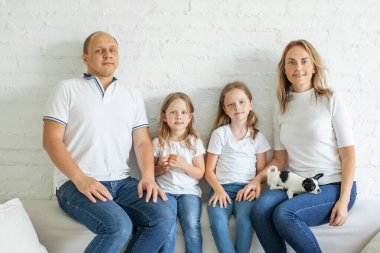 Mutlu anne ve baba ve iki küçük kızı kanepede oturup kameraya bakıyorlar. Beyaz tişört giyen bir aile.