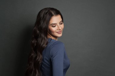 Uzun siyah kıvırcık saçlı, gri arka planda gülümseyen mükemmel kadın modelinin yan görüntüsü.