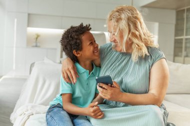 Gülümseyen Afrikalı Amerikalı çocuk ve annesi kanepede oturup, evde rahatlıyor ve akıllı telefonda oynuyorlar.