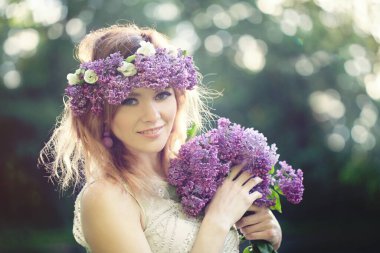 Güzel genç kadın portresi. Leylak çiçekli bahar güzeli çelengi açık havada gülümsüyor.