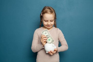 Güzel kız domuz kumbarasına bir dolarlık banknot koyuyor mavi arka plana