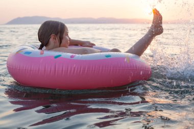 Küçük çocuk pembe şişirilebilir yüzükle gün batımına karşı okyanusta yüzüyor.