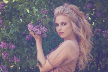 Güzel kadın kameraya bakıyor ve çiçeklerin kokusunu doğayla uyum içinde alıyor.