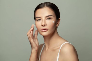 Göz kamaştırıcı, çekici genç bir kadın elinde kremayla yanağına dokunuyor, açık yeşil arka planda izole edilmiş.