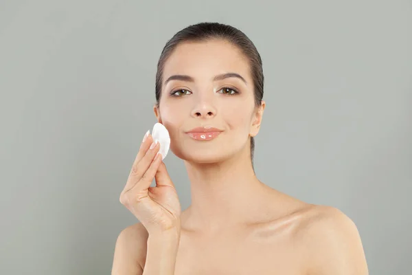 Healthy fresh woman removing makeup from her face with cotton pad. Beauty woman cleaning her face with cotton swab pad isolated on grey background. Skin care and beauty concept.