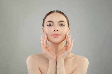 Deri bakımı konsepti. Güzel yüzlü spa modeli sağlıklı yüz portresine dokunuyor. Doğal makyajla gülümseyen güzel bir kadın gri stüdyo arka planında parlayan nemli cilde dokunuyor.