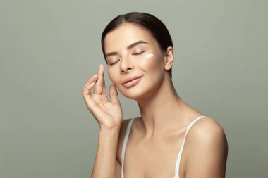 Genç bir kadın yüzüne beyaz krem sürüyor. Gri arka planda kapalı gözlerle gülümsüyor..