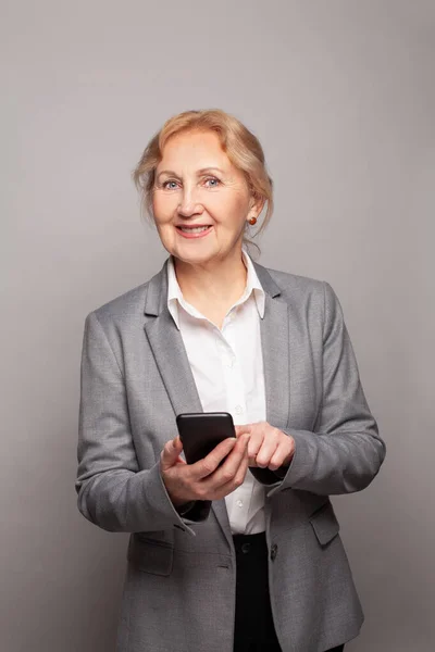 幸せなシニアビジネス女性の肖像グレーのスタジオの壁の背景にスマートフォンを使用してテキストメッセージ — ストック写真