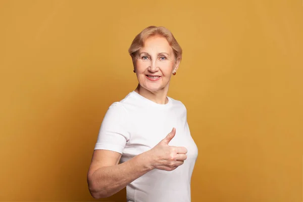 魅力的なシニア女性の肖像60年古いです保持親指アップ上の明るい黄色のスタジオ壁の背景 — ストック写真
