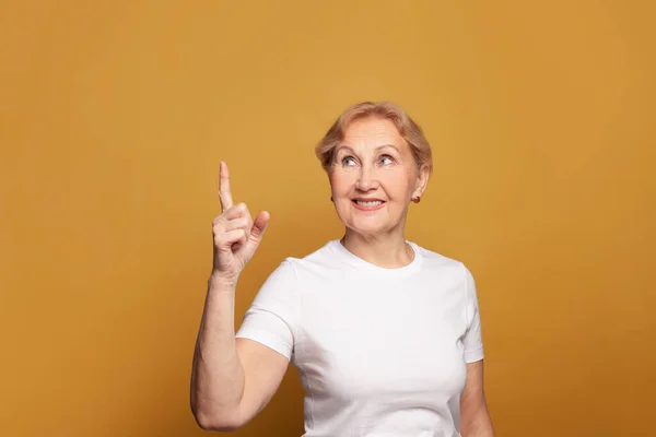 シニア女性で空ホワイトTシャツ指差し指アップ上の明るい黄色スタジオ壁のバナー背景 — ストック写真