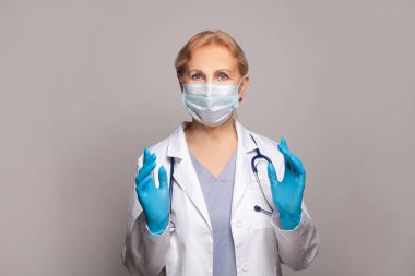 Kadın doktor ayakta duruyor ve gri arka planda jest yapıyor. 