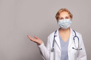 Doktor kadın, gri arka planda tıbbi üniforma giyiyor. Avuç içi kameraya bakıyor ve sunuyor..