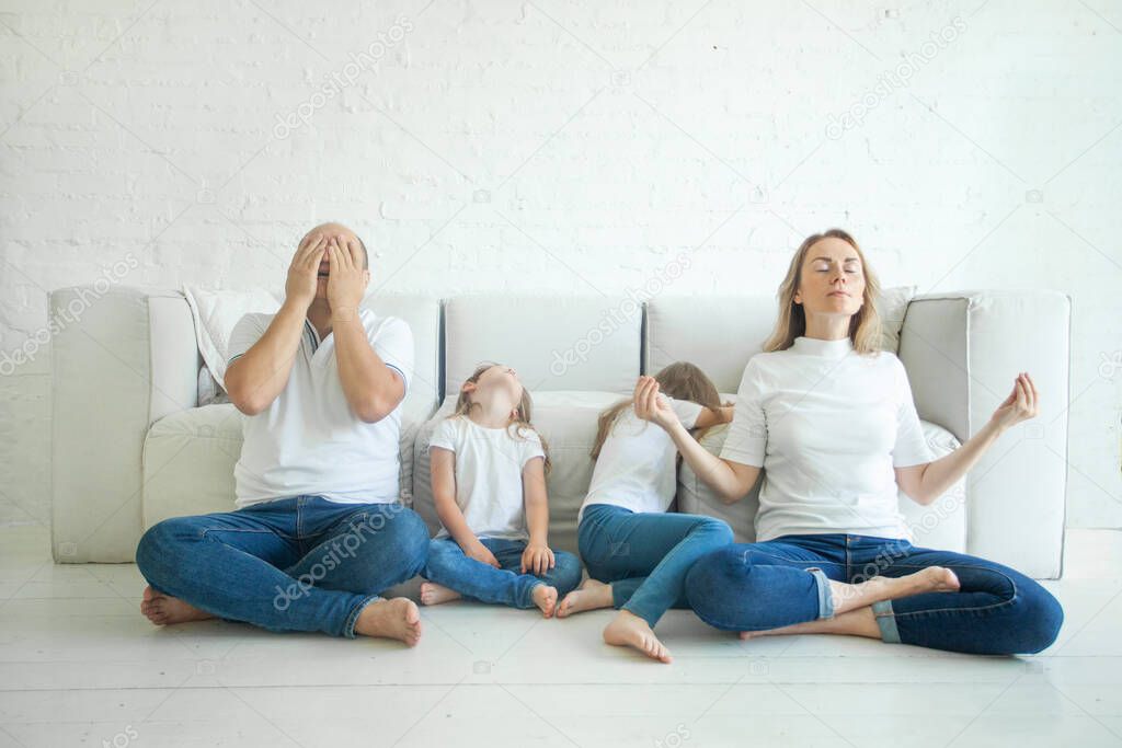 Calm mother with closed eyes meditating in lotus pose on floor, stressed father and two tired children in living room at home.