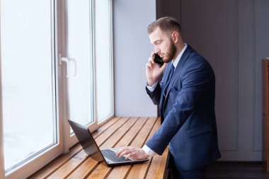 Resmi görevdeki genç işadamı telefonda konuşurken ve ofisteki laptopu kullanarak verileri analiz ederken