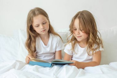 Two kid girl sisters sitting on bed and reading a book at home