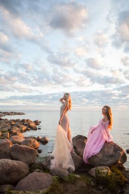 Portrait of two beautiful young gorgeous women wearing fashionable silk dress against stone, sunset sky and clouds