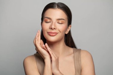 Çekici, genç, spa modeli bir kadın ellerini yüzüne yakın tutuyor ve gri duvar arka planına gülümsüyor, cilt bakımı kavramı.