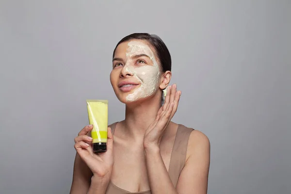 Happy spa model woman with cosmetic mask holding green cosmetic tube box and looking up. Skincare, beauty routine and facial treatment concept