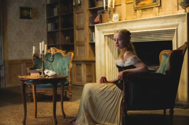 Genç, tatlı, retro tarzı kadın oyuncu eski moda iç mekânlarda koltukta oturuyor.