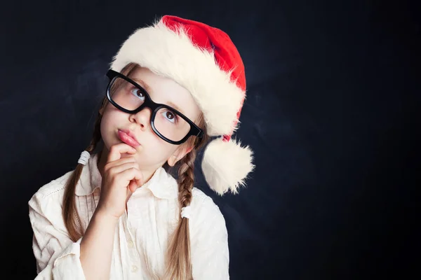 Child in Santa hat thinking. Little girl in Santa hat on chalkboard background with copy space.  Back to school, kid creativity and education concept