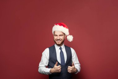 Chtistmas şapkalı neşeli iyimser işadamı gülümsüyor ve kırmızı stüdyo duvarına başparmağını gösteriyor.