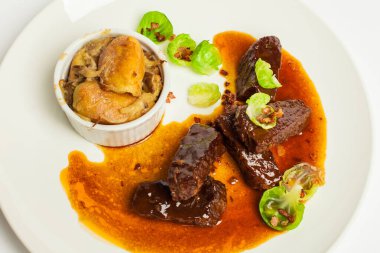 Kırmızı soslu dana yanakları ve yeşil patatesler beyaz arka planda beyaz bir tabakta yakın plan.