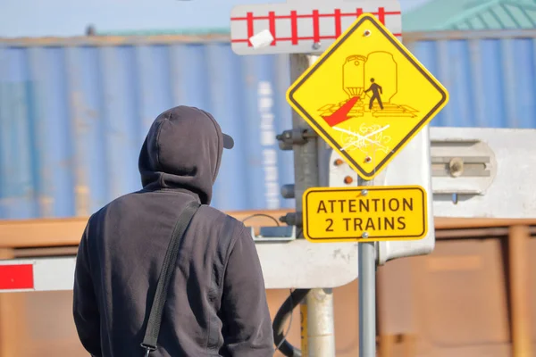 Close, reverse view of a person waiting by a lowered train crossing gate and a sign warning that two trains could be passing.
