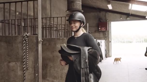 Mulher na engrenagem de equitação transportando sela a cavalo na barraca de estábulos — Vídeo de Stock