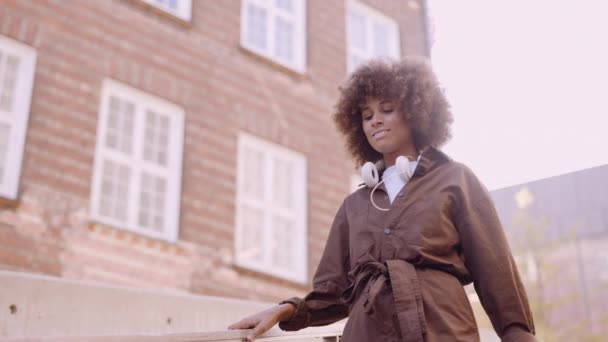 Mujer joven en afro poniéndose auriculares mientras camina — Vídeos de Stock