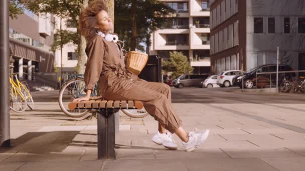 Frau im braunen Overall sitzt auf sonniger Stadtbank — Stockvideo
