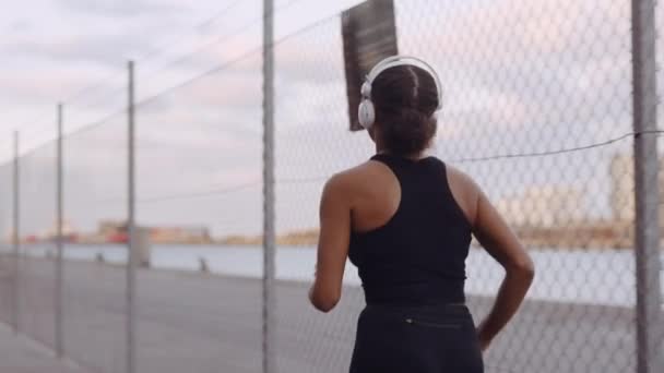 Jeune femme en vêtements de sport et écouteurs jogging le long du port — Video