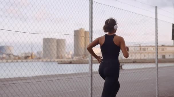 Mujer joven en ropa deportiva y auriculares corriendo a lo largo del puerto — Vídeo de stock