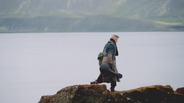Man met gitaarkoffer en grote jas wandelen langs rotsen door Valley River — Stockvideo
