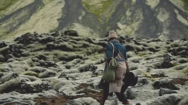 Man wandelen over rotsen in een grote jas met gitaar geval — Stockvideo