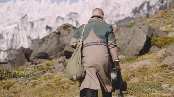 Man in grote jas dragen gitaar geval en zak omhoog heuvel naar gletsjer — Stockvideo