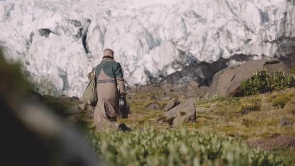 Man i Greatcoat bär gitarr fall och väska upp Hill mot Glacier — Stockvideo
