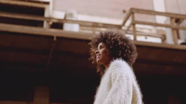 Mujer joven sonriente con pelo afro barrido por el viento caminando por la calle — Vídeos de Stock