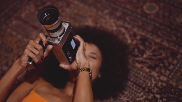 Женщина с афроволосами, лежащая на ковре с помощью винтажной 8-мм камеры — стоковое видео