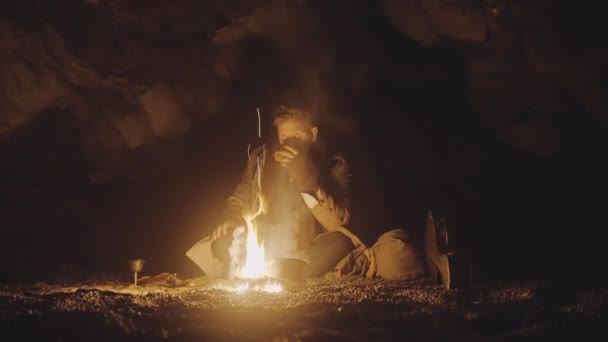Hombre en cueva oscura iluminado por fogata beber de taza — Vídeo de stock