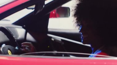 Afro saçlı bir kadın kırmızı arabanın koltuğundan iniyor.